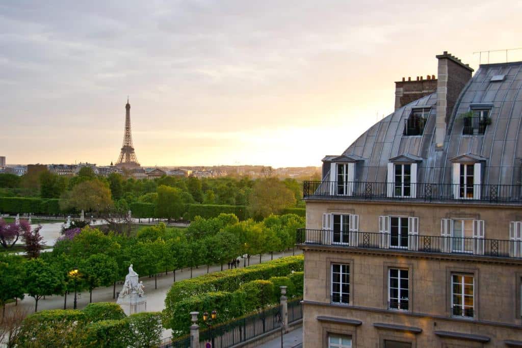 Vista da Torre Eiffel ao pôr do sol, com um parque arborizado e um edifício parisiense clássico em primeiro plano no Hôtel Regina Louvre. Este é um dos hotéis de luxo em Paris.