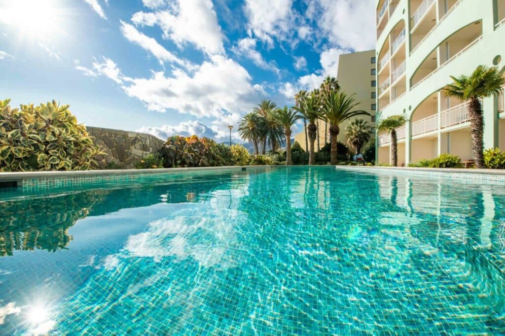Piscina externa ao lado do Pestana Ocean Bay Resort de vários andares do hotel com palmeiras e um céu parcialmente nublado.