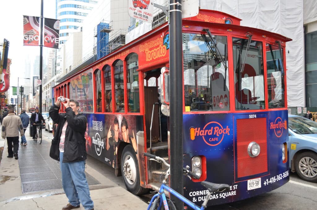 Autocarro vermelho de hop-on hop-off a transportar turistas por ruas movimentadas de Toronto, com fachadas de edifícios modernos ao fundo