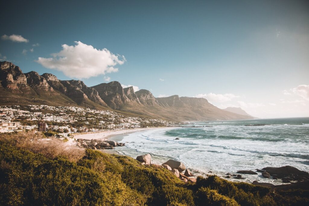 Litoral rochoso de Camps Bay com ondas, praia de areia, vegetação, edifícios e montanhas sob um céu parcialmente nublado.