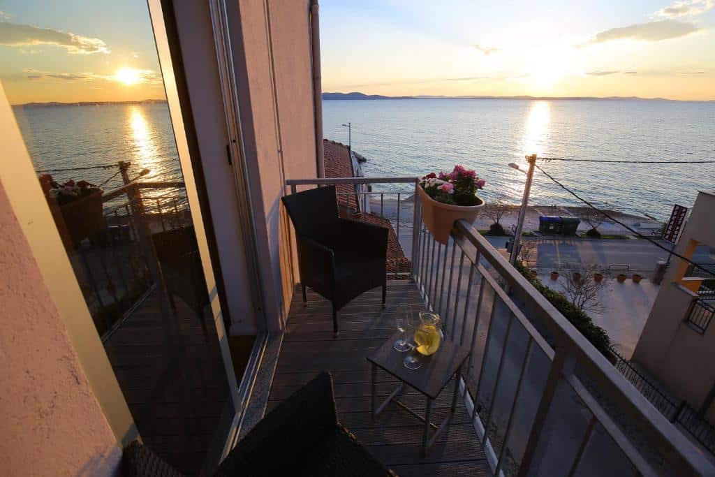 Varanda com vista para o mar ao pôr do sol, cadeiras de vime e mesa com bebidas, no Hotel Delfin, em Diklo