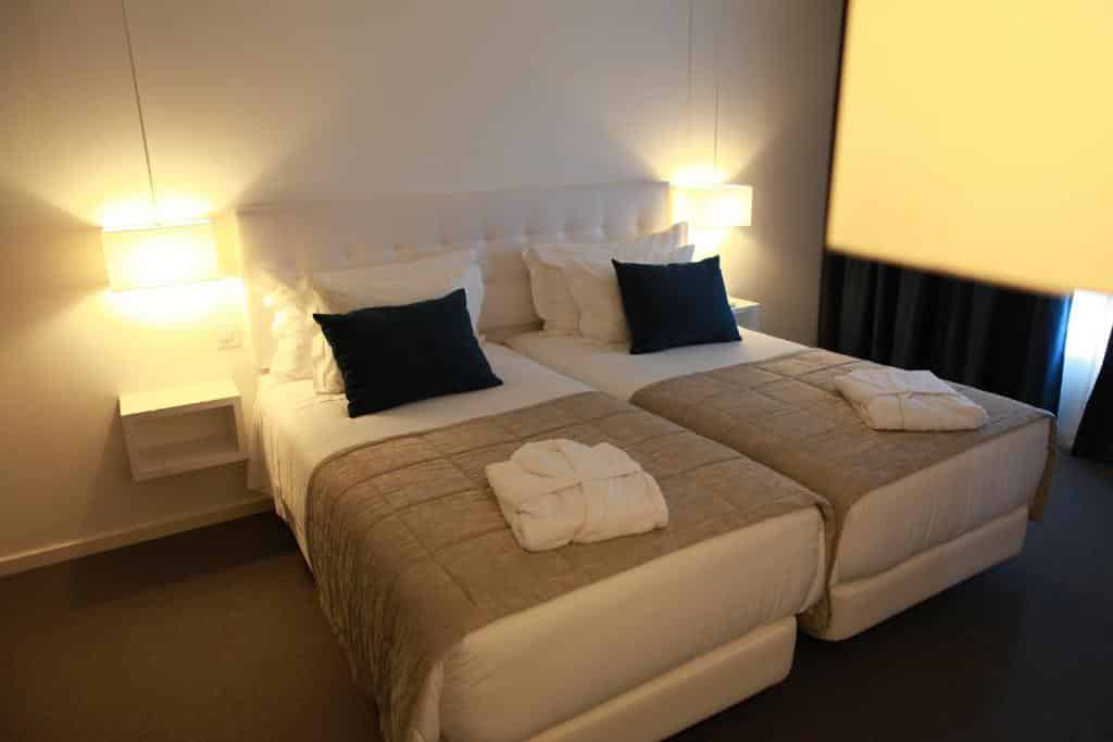 Camas individuais com colchas bege, lençóis brancos, almofadas pretas e roupões dobrados num quarto do Hotel Anjo de Portugal com iluminação suave.