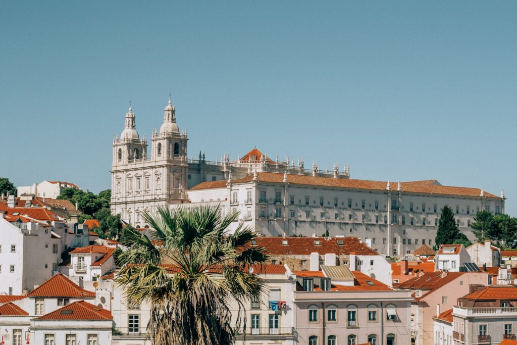 Vista do Mosteiro de São Vicente de Fora, em Alfama, Lisboa: um grande edifício histórico branco com torres ergue-se acima de casas com telhados vermelhos e uma palmeira alta, sob um céu azul claro