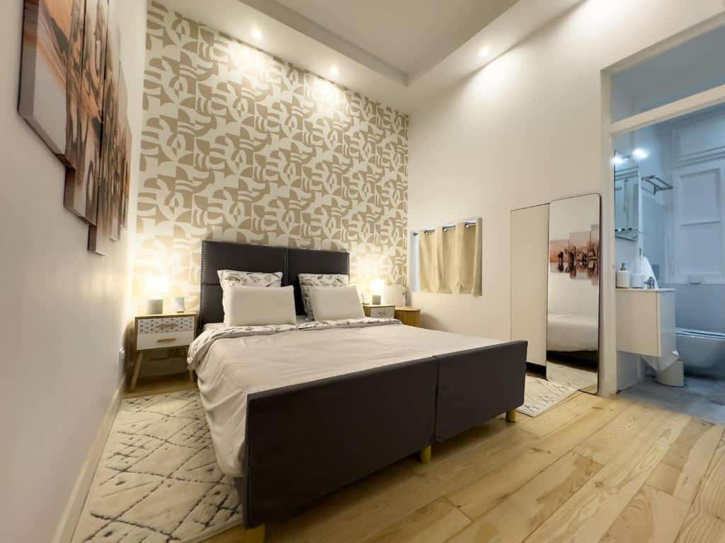 Quarto do Serenity Lisbon Suites com cama de casal, parede com padrão abstrato, tapete, mesas laterais e uma porta aberta que dá para a casa de banho.