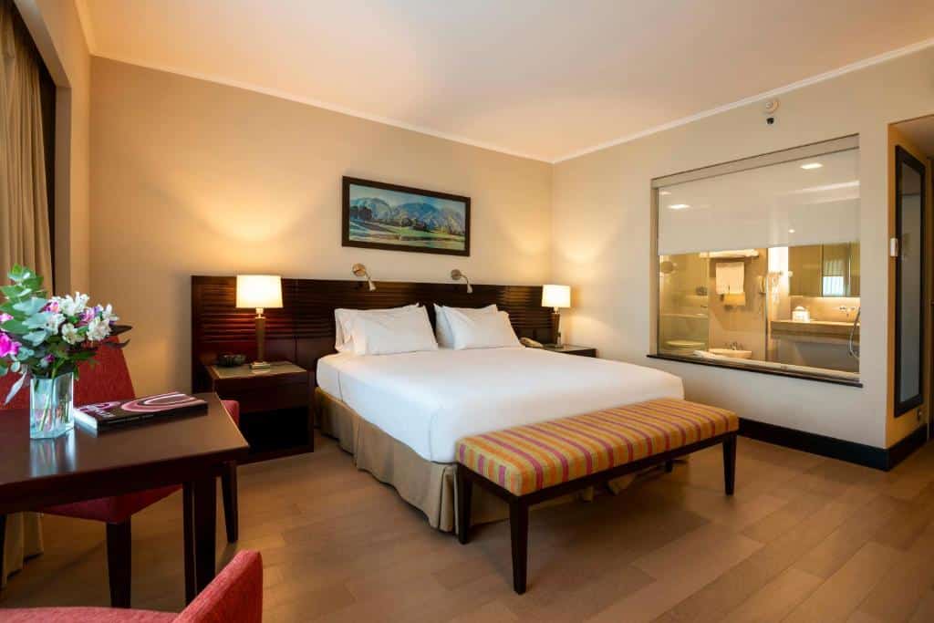 quarto do Eurobuilding Hotel Boutique Buenos Aires com cama centralizada, mesas e candeeiros dos dois lados, mesa de madeira com poltronas vermelhas e uma casa de banho com cortina retrátil