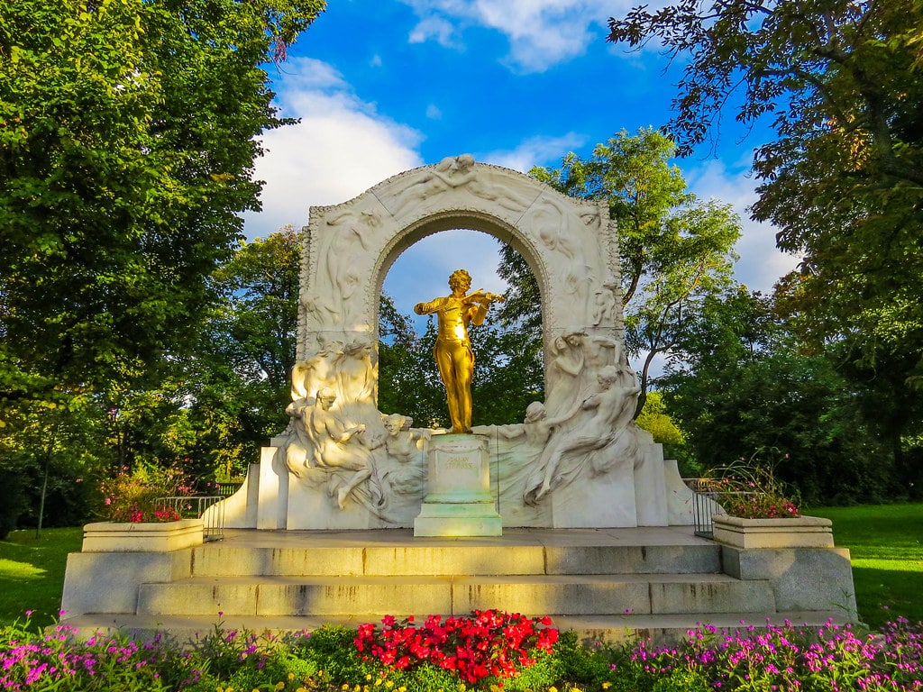 Monumento dourado a Johann Strauss no Stadtpark, com a estátua de um violinista sob um arco decorativo, num parque arborizado.