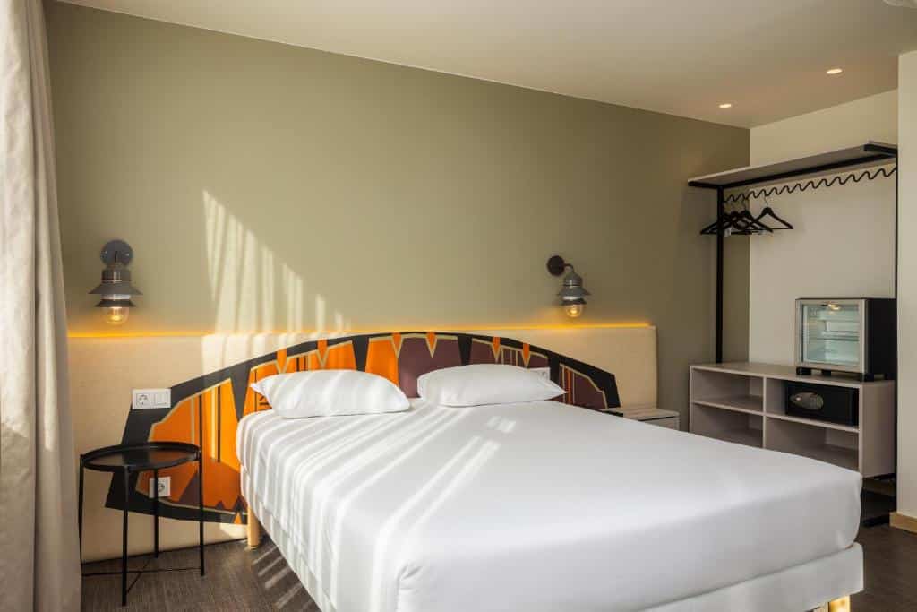 Quarto do Hotel Ibis Styles Lisboa Liberdade com uma cama de casal e um pequeno frigorífico junto a um armário de conceito aberto com bengaleiro