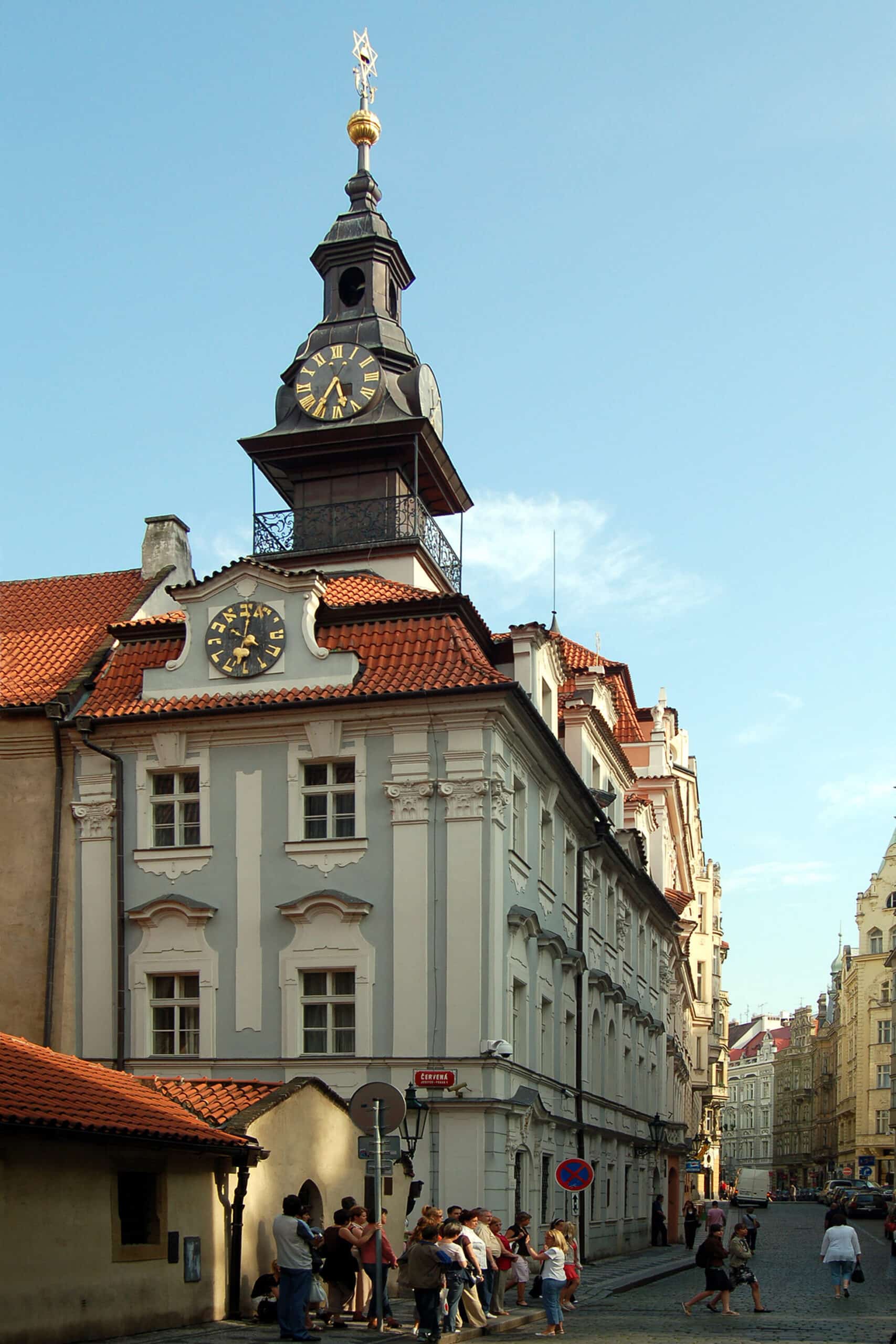 Edifício histórico com uma alta torre de relógio numa cidade europeia; pessoas reúnem-se e caminham pela rua de pedra em baixo.
