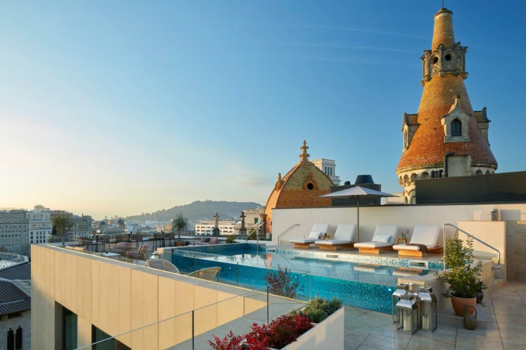 Piscina do ME Barcelona, com espreguiçadeiras e um guarda-sol aberto, e vista sobre a cidade ao fundo.