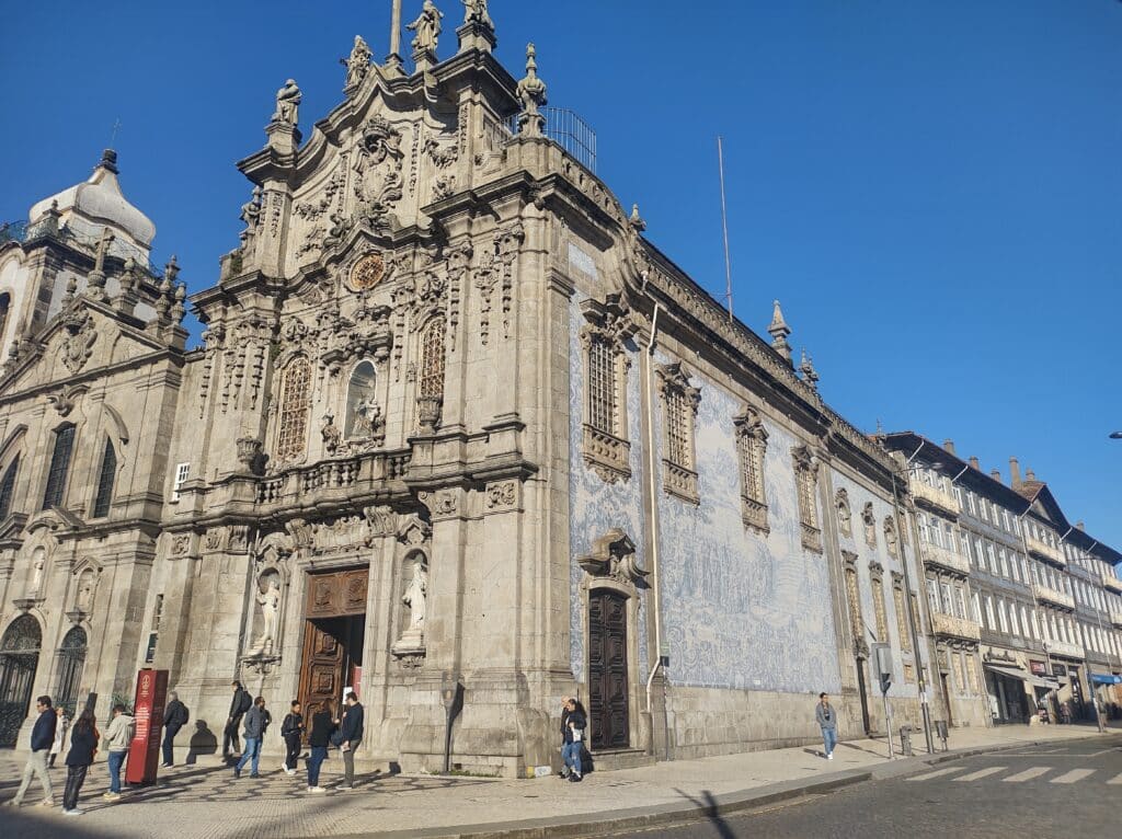 Pessoas passam junto a uma grande igreja de pedra ornamentada, com painéis de azulejos azuis e brancos, num dia soalheiro: a Igreja do Carmo, no Porto.