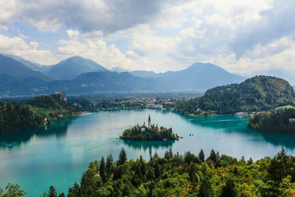 Vista panorâmica do Lago Bled, na Eslovénia, famoso pela água azul-turquesa e por uma pequena ilha com uma igreja no centro. É um dos destinos invulgares da Europa. Ao fundo, montanhas cobertas de florestas e nuvens dispersas no céu completam a paisagem. O ambiente é sereno e pitoresco, destacando a beleza natural e histórica do local.