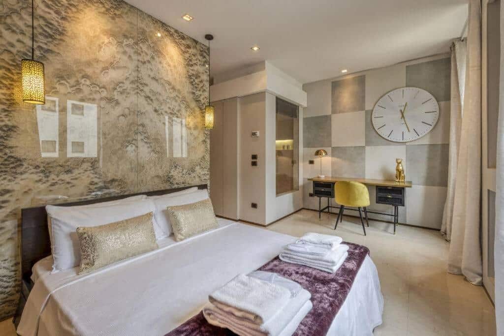 Quarto do Milan Royal Suites – Centro com uma cama de casal com almofadas e toalhas, dois candeeiros suspensos, uma secretária com uma cadeira e uma varanda com cortinas