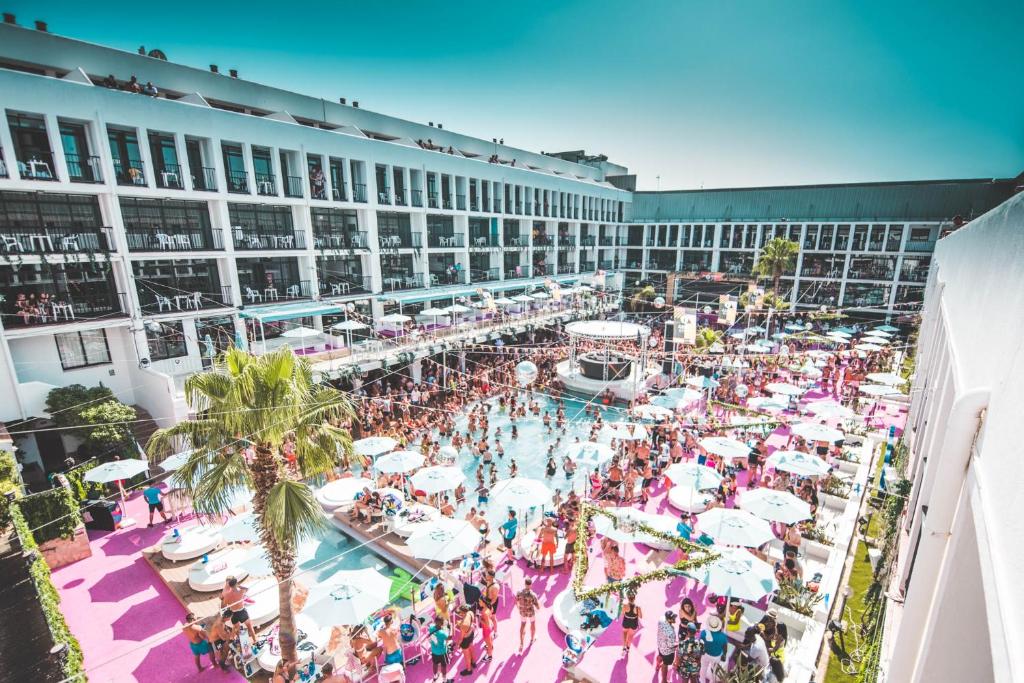 Grande festa na piscina exterior do Ibiza Rocks Hotel, com muita gente, chão cor-de-rosa, chapéus de sol e varandas com vista para o local.