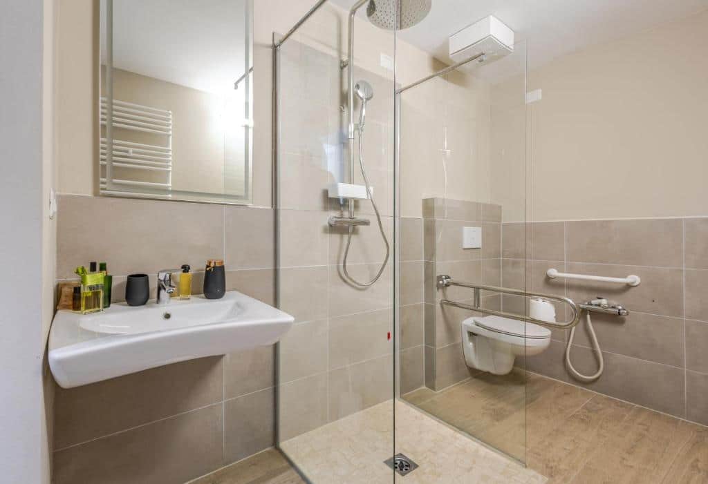 Casa de banho acessível com base de duche em vidro, lavatório branco e barras de apoio