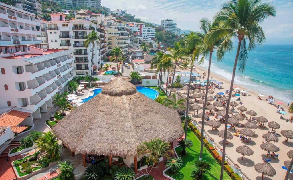 Um resort à beira-mar com uma grande cabana de telhado de colmo, piscina, palmeiras e chapéus de sol ao longo da costa arenosa.