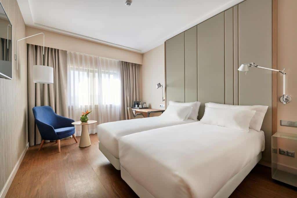 Quarto do NH Lisboa Campo Grande com duas camas individuais, uma janela ampla com cortinas, uma pequena mesa com utensílios para café, uma poltrona e uma televisão em frente às camas.
