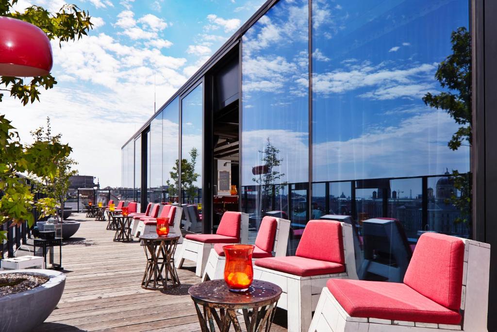 Lounge ao ar livre na cobertura do 25hours Hotel, com assentos de almofadas vermelhas, mesas pequenas e paredes de vidro que refletem o céu azul.