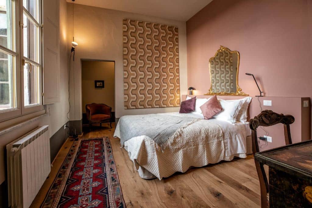 Quarto da Ca’ Bianca, com parede cor de salmão, cama de casal com almofadas, tapete no chão de madeira, mesa de madeira com cadeira e um outro espaço com uma poltrona estofada de época