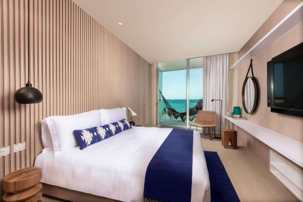 Quarto no SLS Cancun Hotel & Spa com cama de casal, varanda virada para o mar com uma rede, espelho, televisão e poltrona, decorado em tons de branco e azul