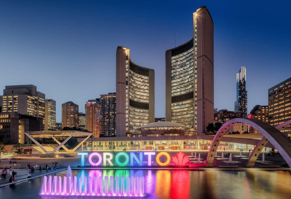 Skyline de Toronto, Canadá, com edifícios modernos de escritórios e centro de convenções iluminados à noite, com fonte ornamental e letreiro colorido com o nome da cidade em primeiro plano