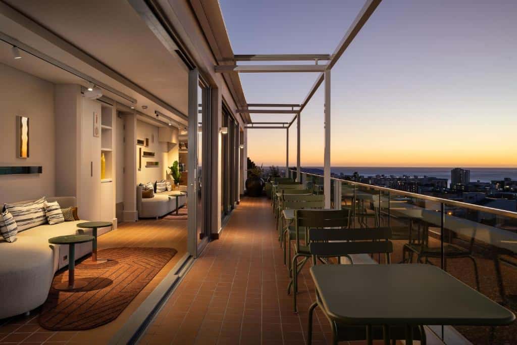 Terraço ao ar livre no Home Suite Hotels Sea Point com mesas e cadeiras ao pôr do sol, com vista para a cidade e o oceano.