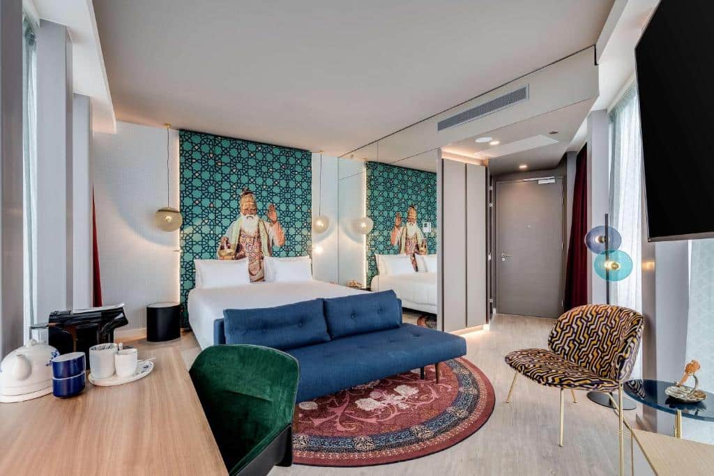 Quarto do nhow Amsterdam Rai, com cama de casal, sofá azul sobre um tapete estampado e uma zona de café com cadeira verde
