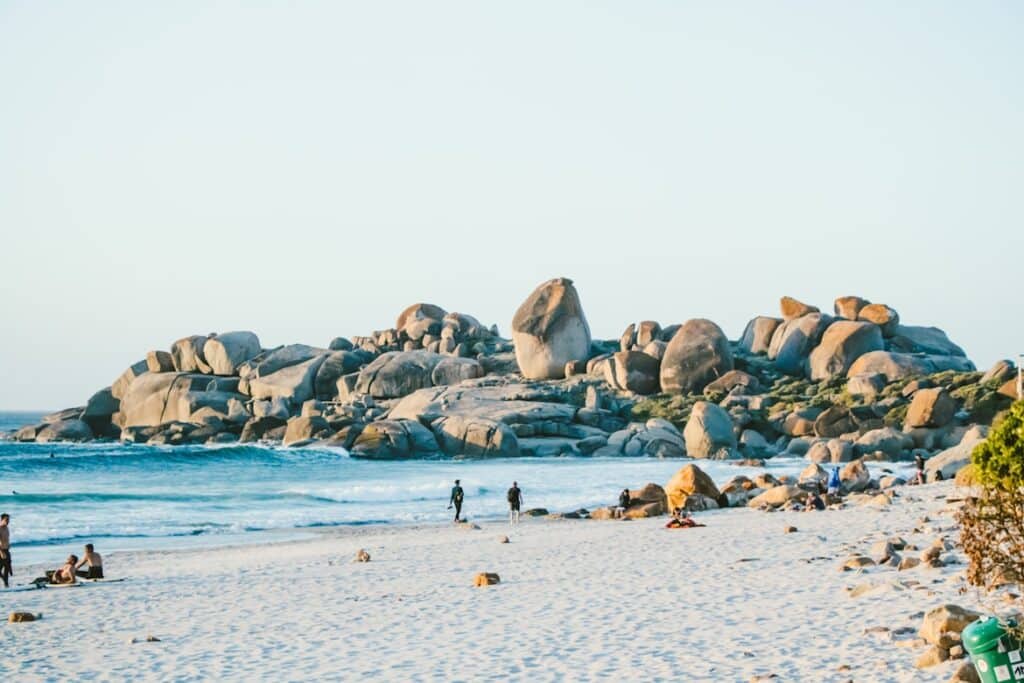 Praia de Llandudno, na Cidade do Cabo, com pessoas a caminhar junto a grandes formações rochosas à beira-mar sob um céu limpo.