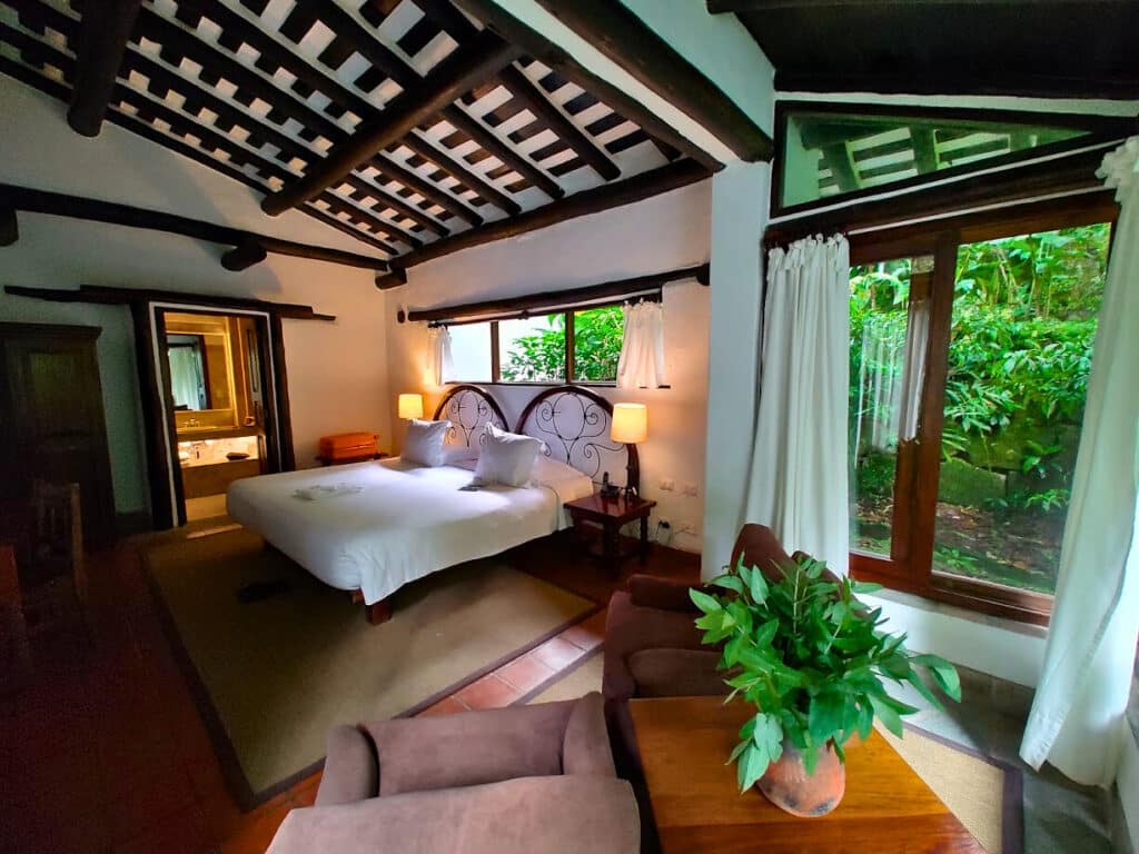 Quarto do Inkaterra Machu Picchu Pueblo Hotel com cama de casal, área de estar com poltronas e ampla janela com vista para a vegetação