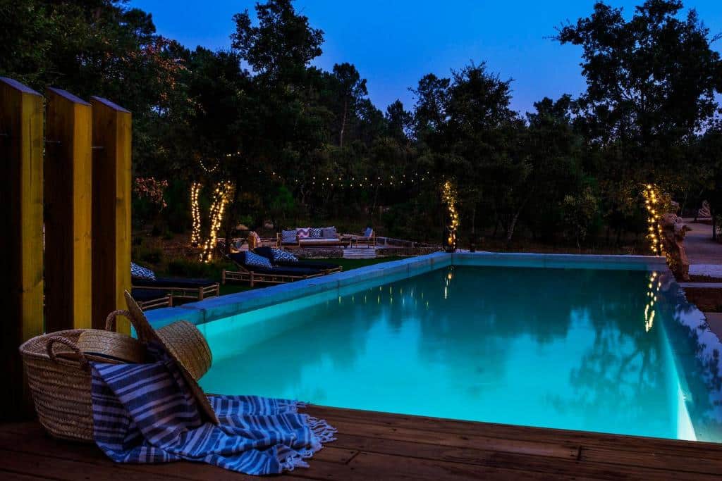 Piscina ao anoitecer com luzes brilhantes, cestos de vime, uma toalha e lugares de lounge ao fundo entre as árvores, no alojamento Luz Charming Houses.