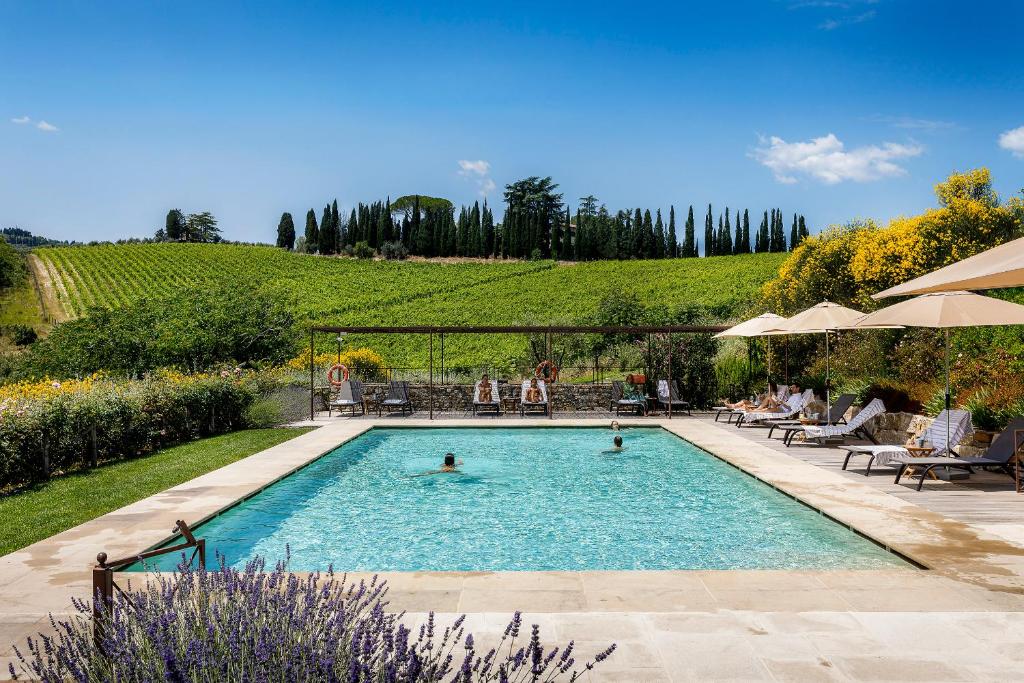 Piscina retangular grande do Borgo Del Cabreo, um dos hotéis na Toscana, com espreguiçadeiras à volta e vista sobre o amplo campo verde do local, com vinhas, árvores e flores lilás e amarelas