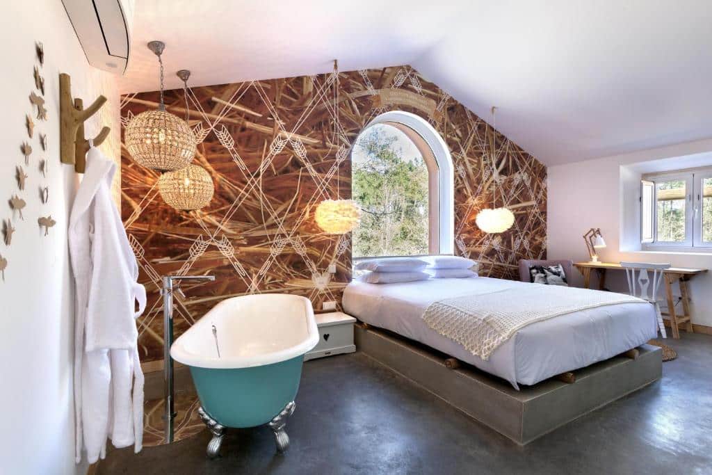 Quarto moderno com uma grande janela, uma cama de casal, banheira com pés de garra em azul-petróleo, secretária e parede de destaque com padrão de palha, no Luz Charming Houses, para representar onde ficar em Fátima.