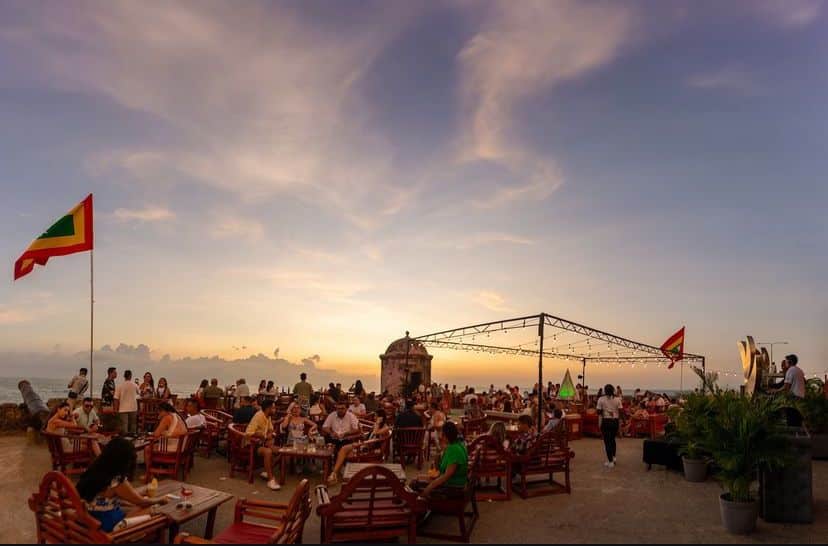 Esplanada ao ar livre do Café del Mar, em Cartagena, com mesas de madeira num terraço virado para o mar ao pôr do sol.