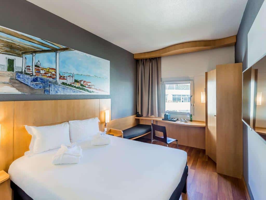Quarto do Hotel Ibis Lisboa Parque das Nações com cama de casal, lençóis brancos, mobiliário de madeira e secretária com cadeira.