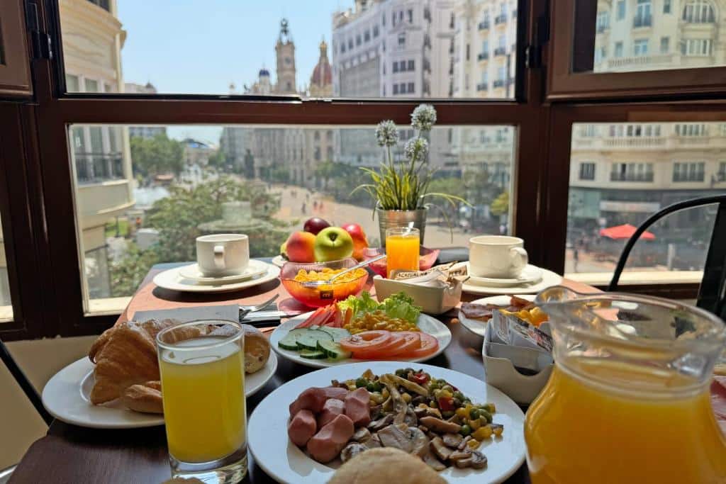 Pequeno-almoço do Casual Vintage Valencia com vista panorâmica da cidade, com pratos de fruta, sumo de laranja, pães e frios numa mesa de decoração floral diante das janelas viradas para os edifícios do centro.