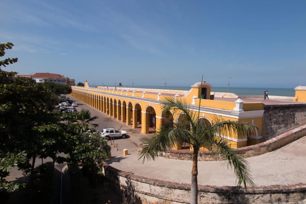 Longo edifício colonial amarelo de Las Bóvedas, em Cartagena, com uma sucessão de arcos, junto ao mar e rodeado de palmeiras.