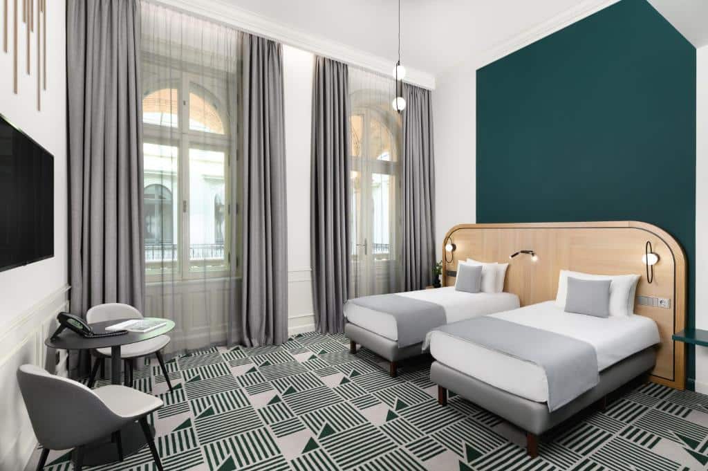 Quarto do Hotel Oktogon Haggenmacher com duas camas de solteiro, uma mesa com duas cadeiras, janelas grandes com cortinas e alcatifa com padrão geométrico.