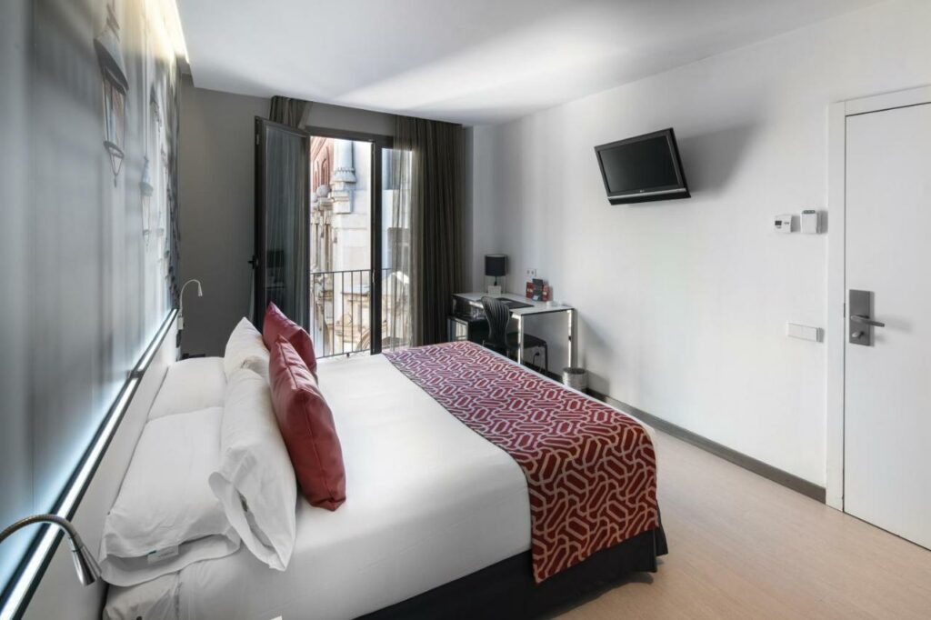 Quarto do Catalonia Avinyo. A cama de casal está virada para uma televisão e tem uma secretária com candeeiro junto à porta de vidro que dá para a varanda.