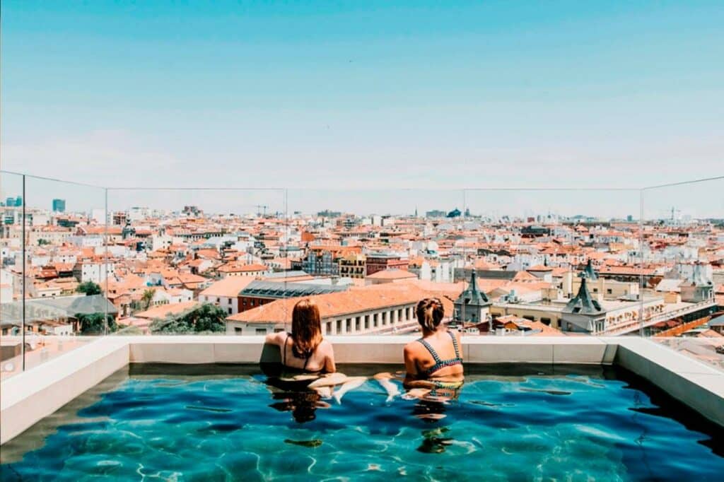 Duas mulheres sentadas dentro da piscina do Dear Hotel Madrid a observar a vista da cidade. Estão de costas para a fotografia, com apenas a parte de cima do corpo fora de água. A fotografia foi tirada de dia e há proteção de vidro à volta da piscina.