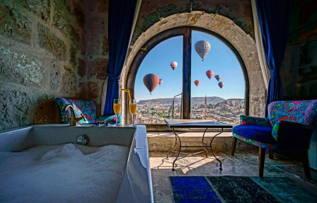 Sala do Arinna Cappadocia, um dos hotéis da Capadócia, com paredes de pedra, banheira, duas cadeiras, uma mesa e balões de ar quente visíveis através de uma grande janela em arco.
