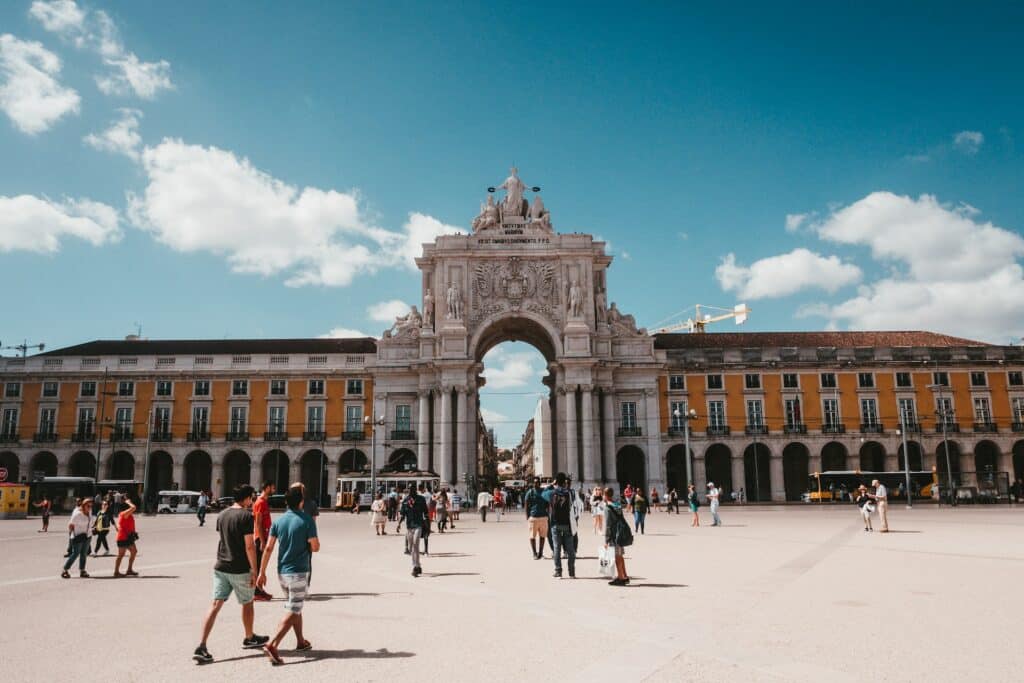 Praça do Comércio em Lisboa com um grande arco com esculturas, edifícios antigos em tons de cinzento e laranja e muitas pessoas a caminhar pelo local
