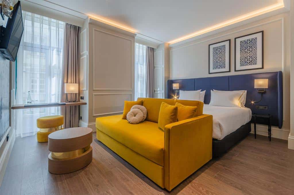 Quarto do Eurostars Lisboa Baixa com uma cama de casal e um sofá aos pés, duas varandas com cortinas, dois pufes e uma pequena bancada com um candeeiro
