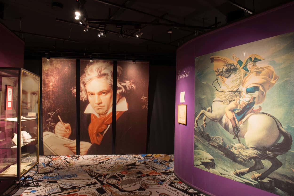 Exposição na Haus der Musik, com grandes retratos de Beethoven e Napoleão, rodeada por paredes roxas e piso estampado.