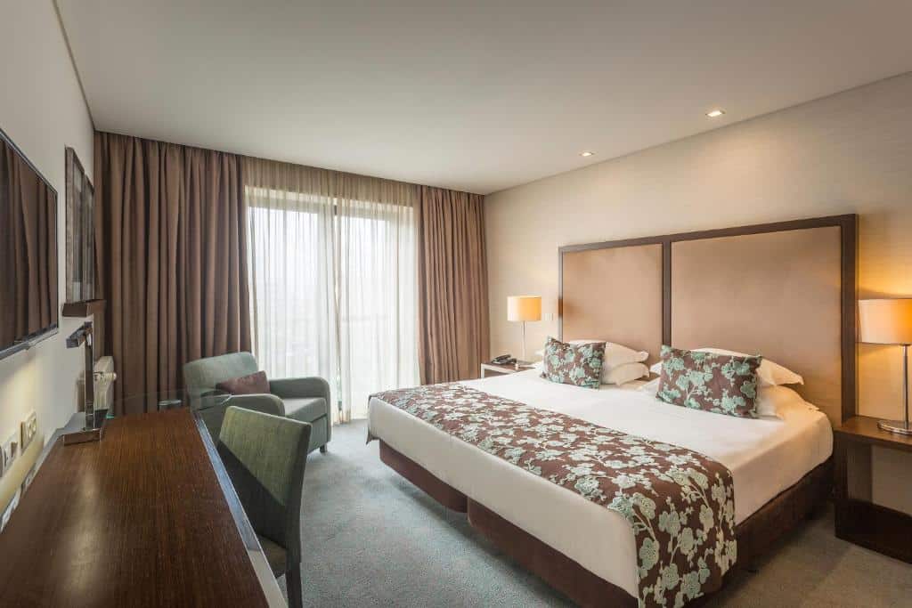 Quarto no Hotel Dom Gonçalo & Spa com cama de casal grande, poltrona, secretária, TV, candeeiros e cortinados até ao chão a cobrir uma janela.