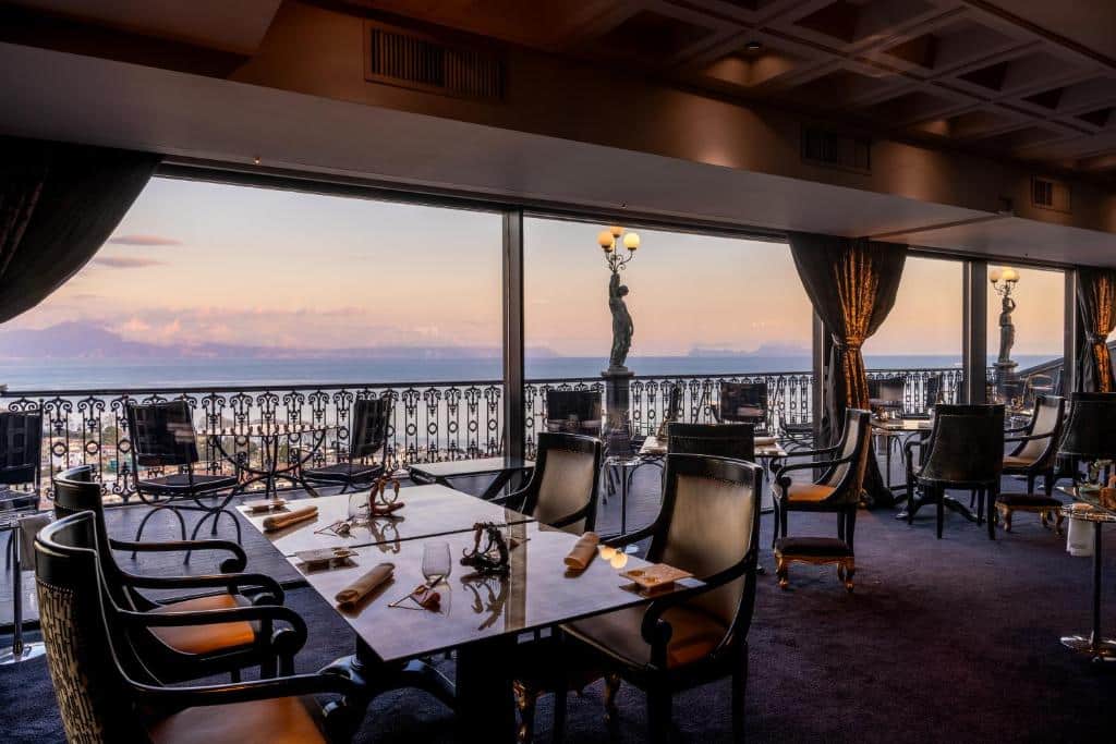 Elegante sala de jantar do Grand Hotel Parker's com vista panorâmica sobre a cidade ao pôr do sol, mesas e cadeiras de estilo clássico, candeeiros e pormenores decorativos em metal.