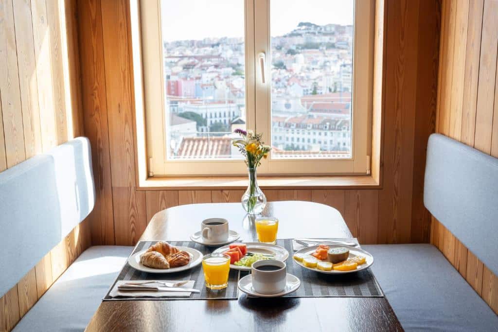 Pequeno-almoço com café, sumo, bolos e fruta servido numa mesa junto a uma janela com vista para a cidade, no Lisboa Pessoa Hotel.