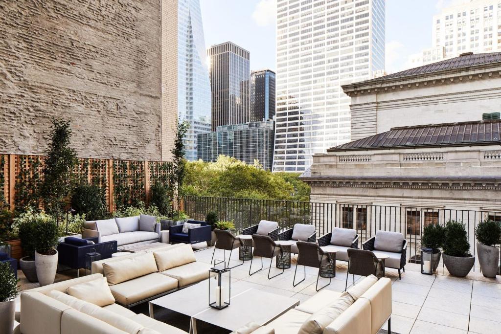 No terraço do Park Terrace Hotel há um sofá de três lugares em primeiro plano e uma mesa de centro com um candeeiro. À direita, um sofá de dois lugares. Junto à parede de tijolo, várias plantas em vasos e, ao longo da grade, uma fila de cadeiras voltadas para a vista dos edifícios de Nova York.
