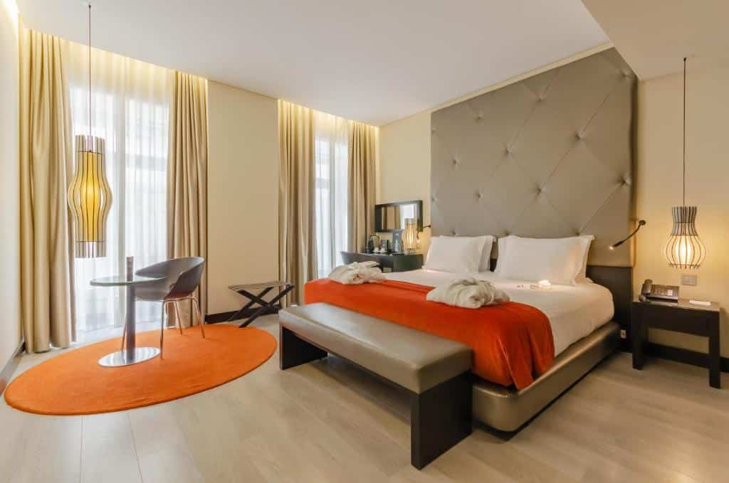 Quarto do Hotel Santa Justa com cama de casal e cabeceira estofada, uma janela com cortinas à esquerda, uma mesa com serviço de café e duas cadeiras, e mesas de cabeceira com candeeiros.