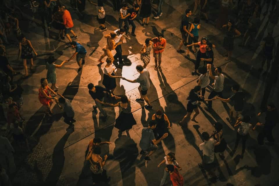 Vista de cima de pessoas a dançar salsa energicamente num espaço aberto com pouca luz, projetando longas sombras no chão.