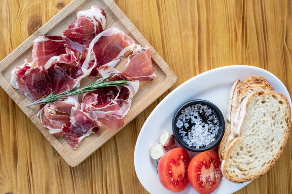 Fatias de presunto ibérico numa tábua, ao lado de um prato com pão, tomate cortado ao meio e um recipiente com sal grosso.