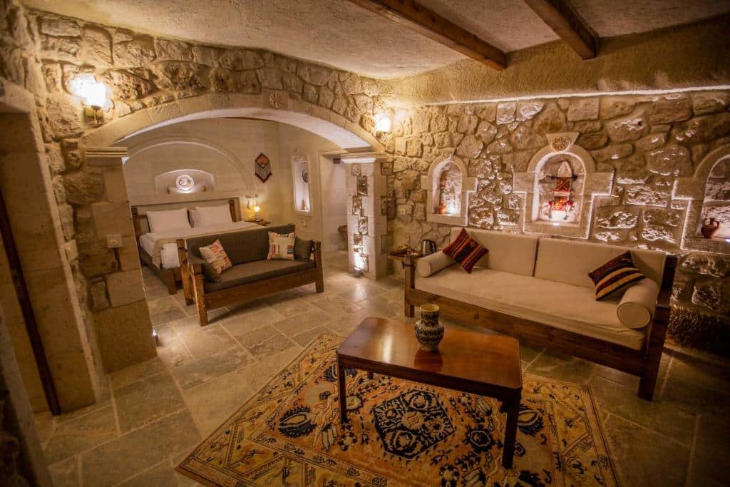 Sala de estar do Zara Cave Hotel, um dos hotéis da Capadócia, com paredes de pedra, dois sofás, uma cama, mesa de centro de madeira e um tapete estampado sobre piso de azulejo.